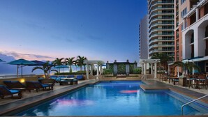 Zona de piscina con sillas de descanso y sombrillas, rodeada por un edificio de gran altura.