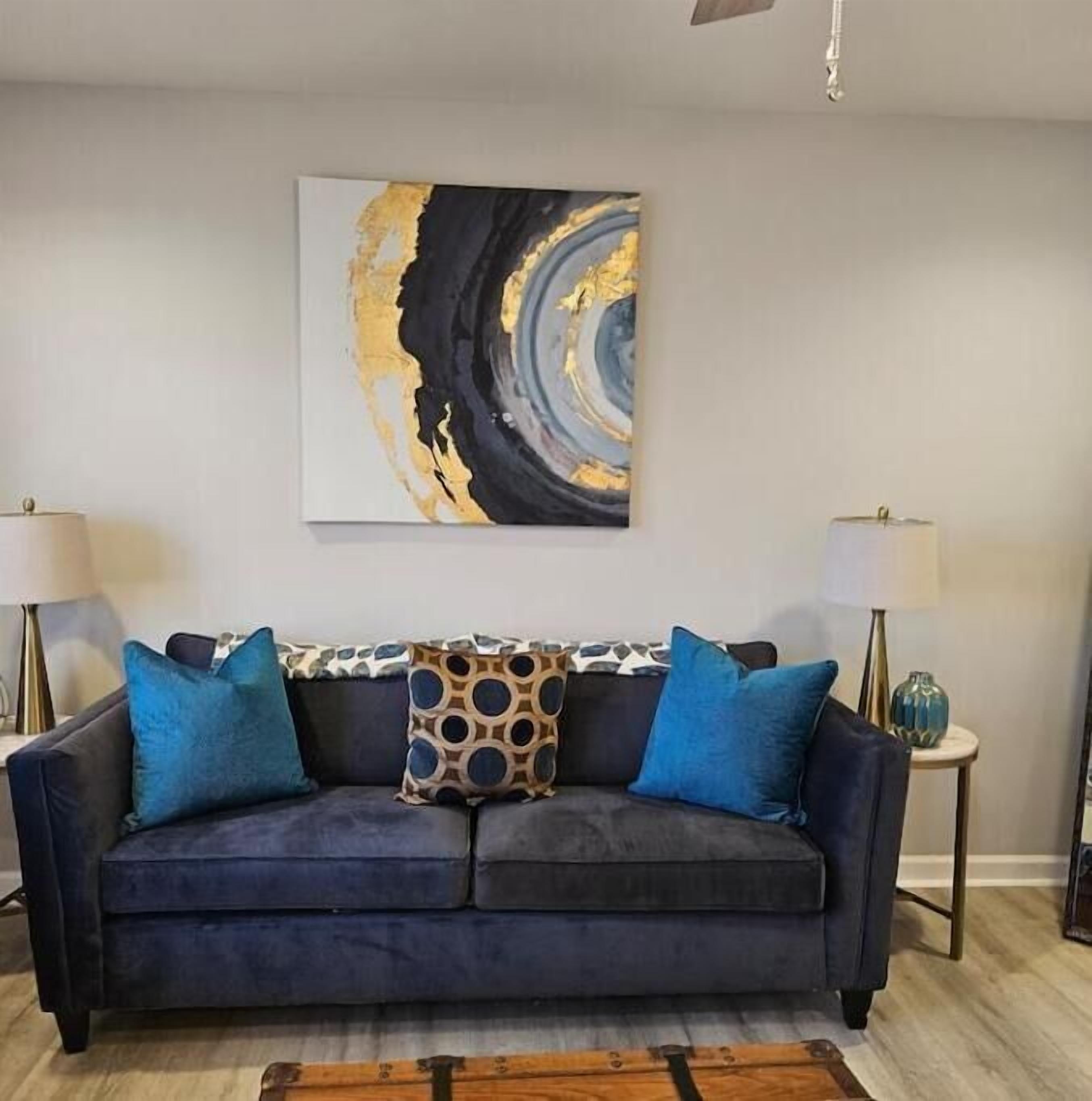 Un salon avec un canapé bleu foncé, deux coussins bleus, un coussin décoratif à motifs et un grand tableau abstrait accroché au mur. Deux lampadaires sont placés de part et d’autre du canapé.