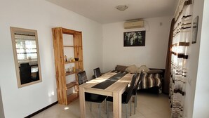 Un coin repas avec une longue table, des chaises, une bibliothèque en bois, un lit avec une literie à rayures et une fenêtre avec des stores.