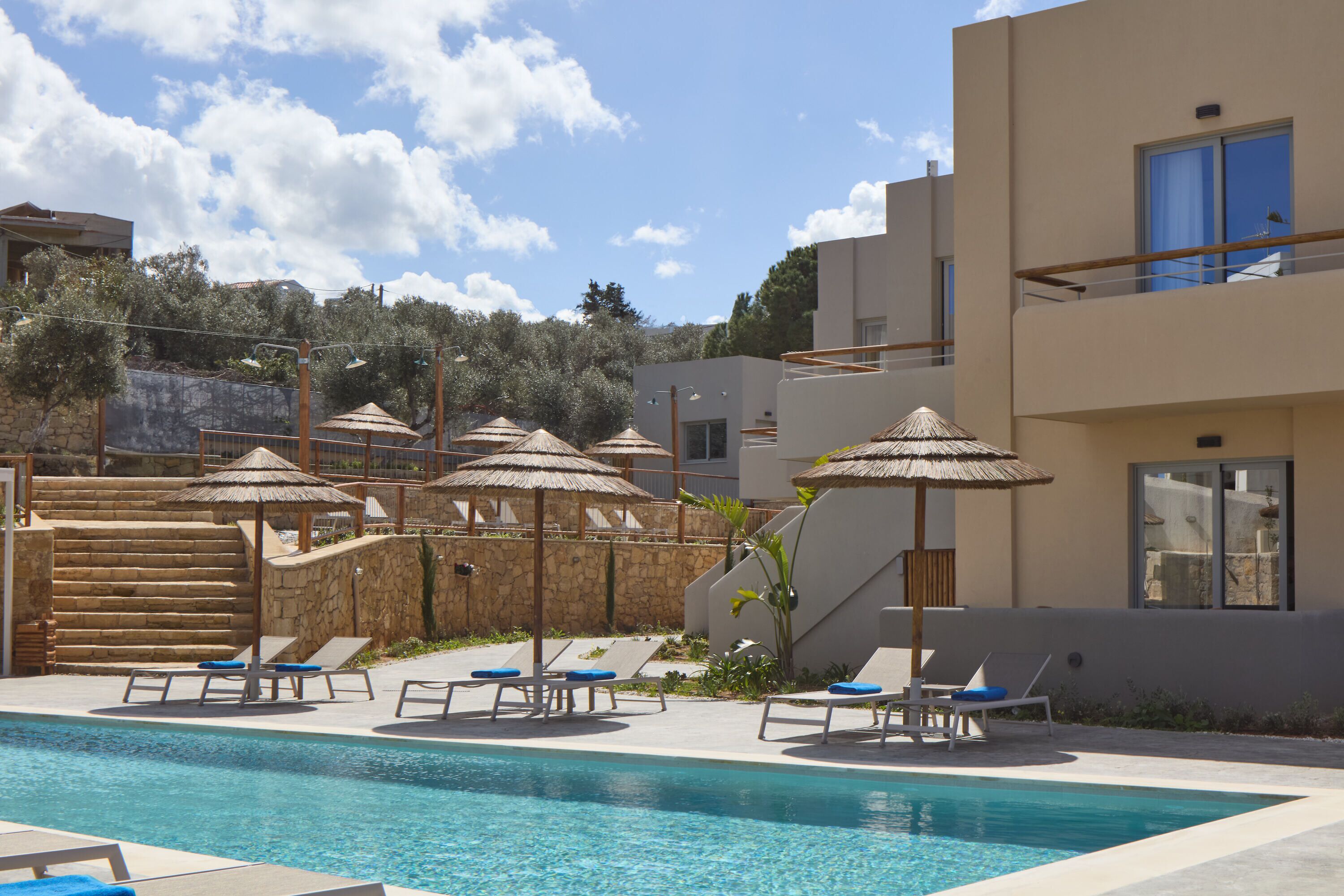 A swimming pool with lounge chairs and thatched umbrellas, surrounded by modern buildings and a stone wall.