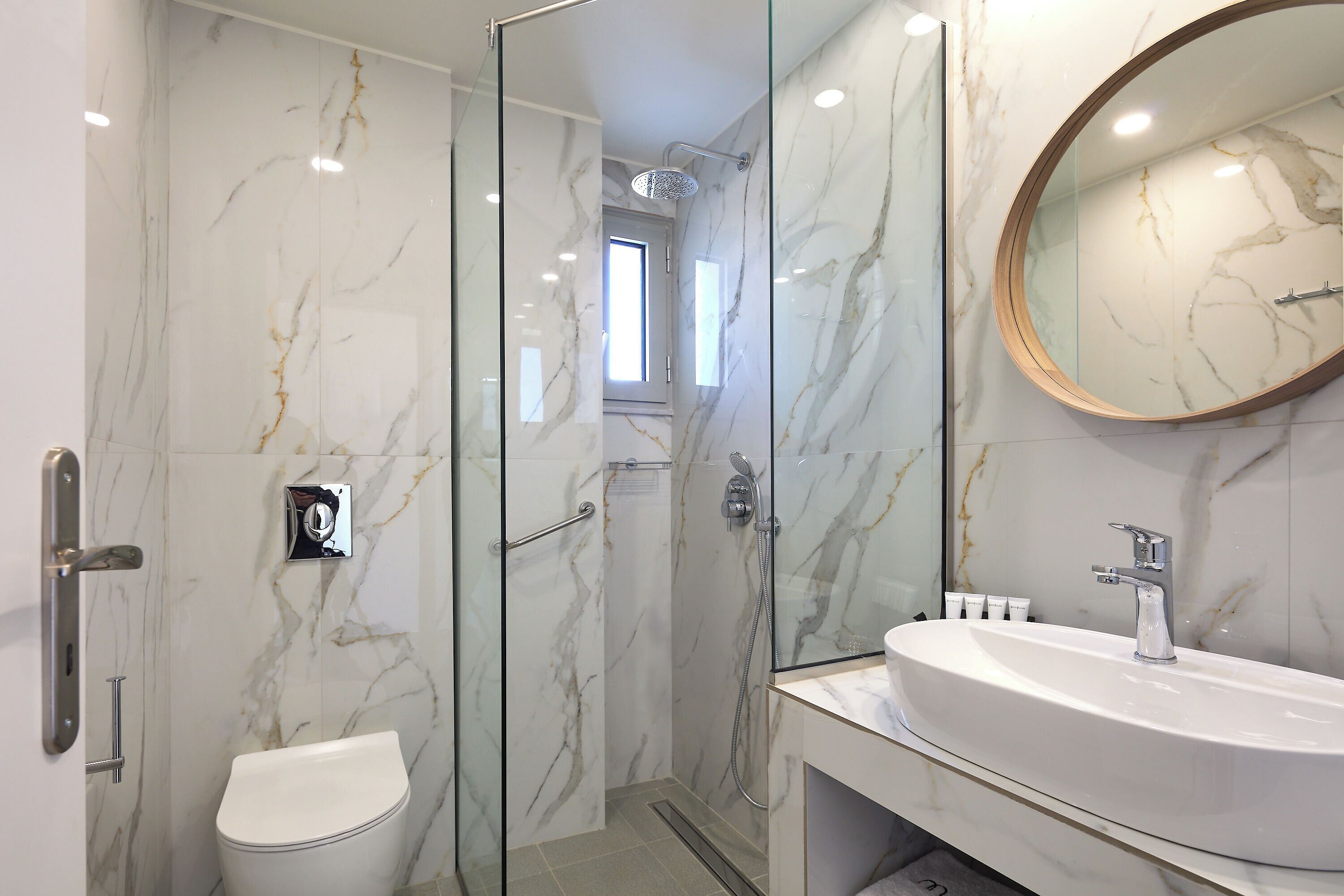 Une salle de bain moderne avec une douche à parois de verre, un mur en marbre, un miroir rond et un lavabo blanc.