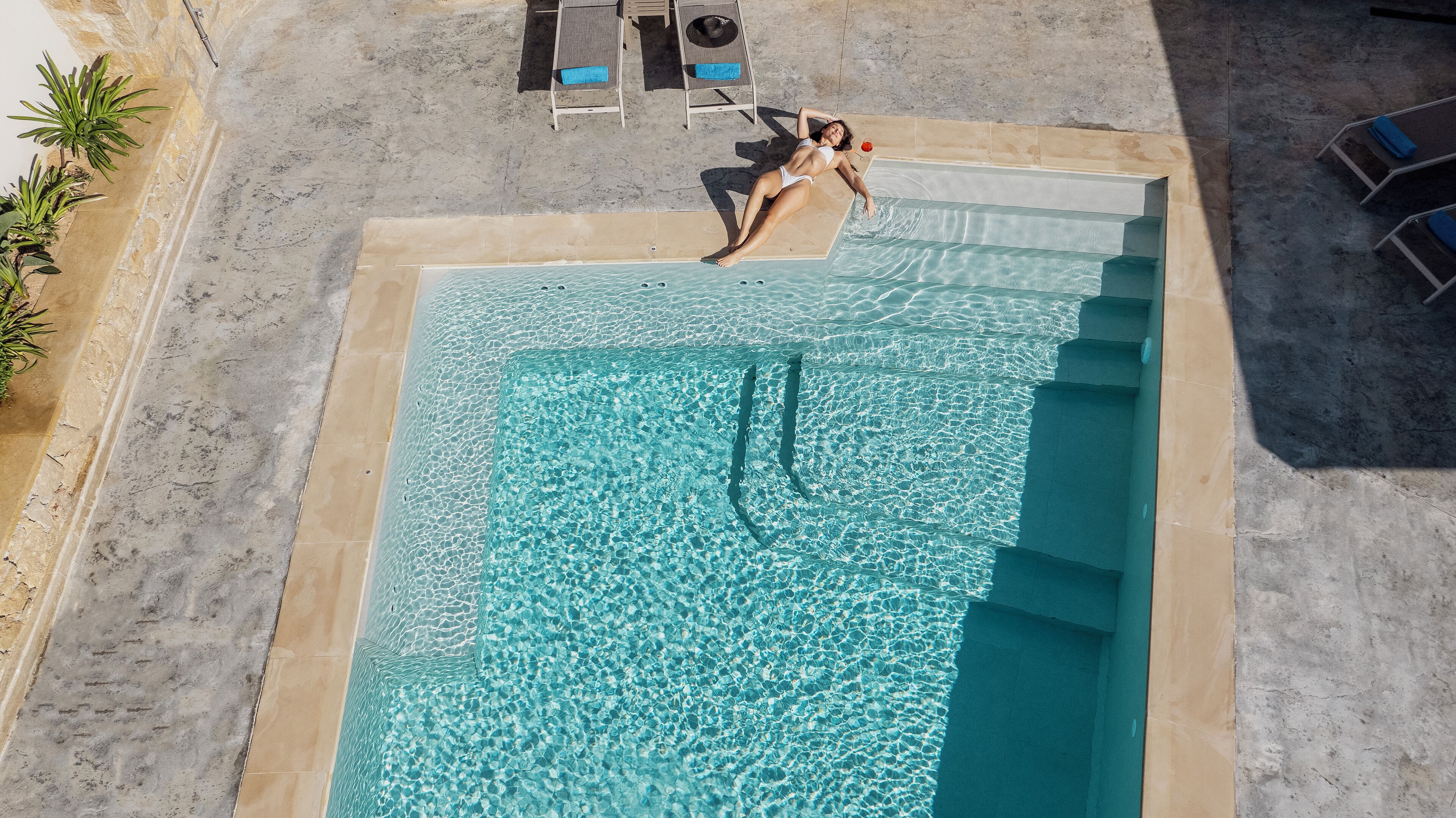 Vue aérienne d’une piscine rectangulaire à l’eau d’un bleu limpide, entourée d’une terrasse carrelée et de chaises longues.