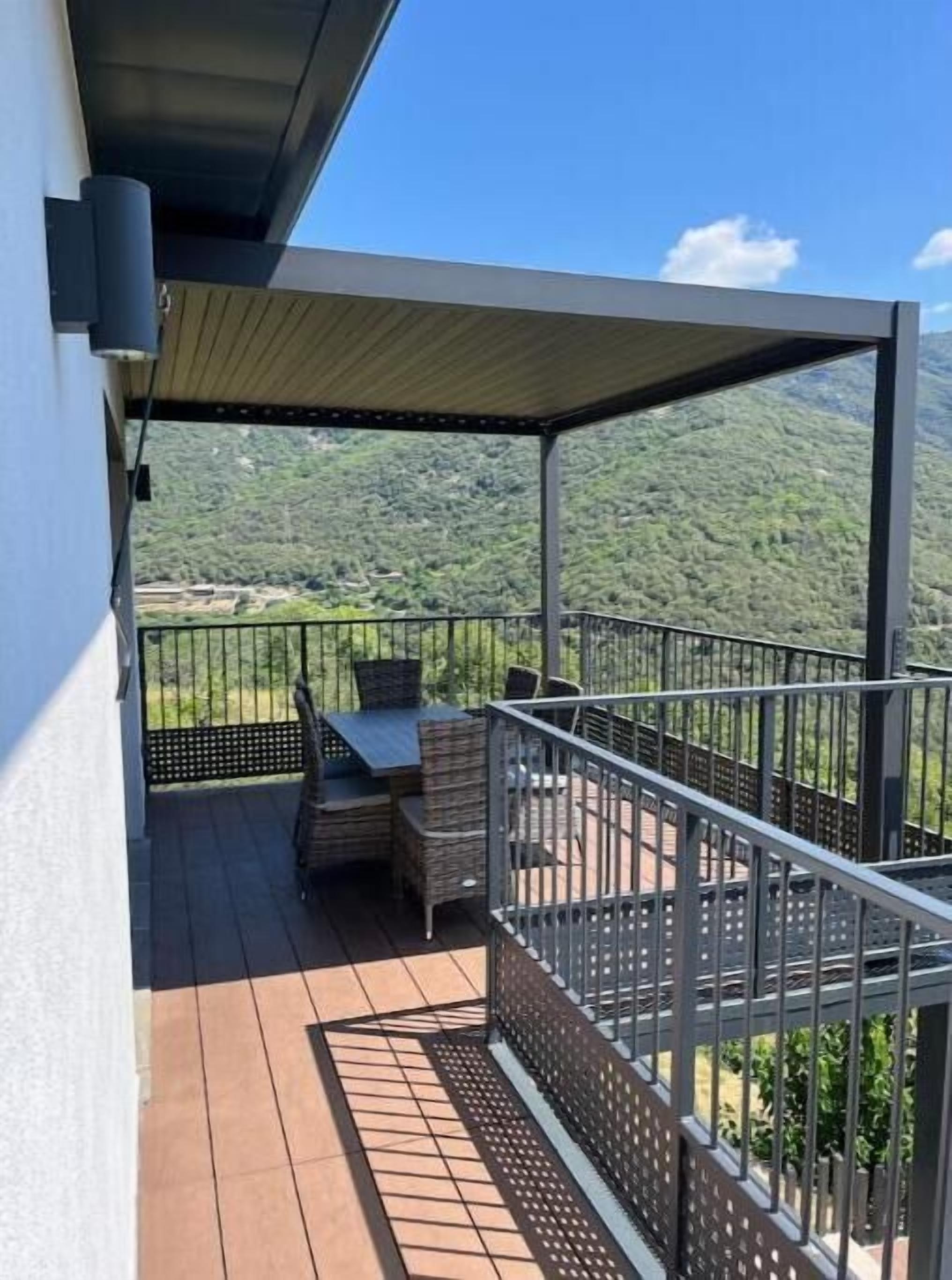Un balcón techado al aire libre con una mesa y sillas, con vistas a un paisaje montañoso.