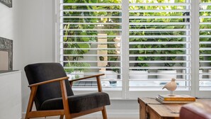 A room with a black armchair, a wooden side table with a book, and a view through white blinds to a garden. - Villa Marguerite - walking distance to the beach (Coolum Beach)