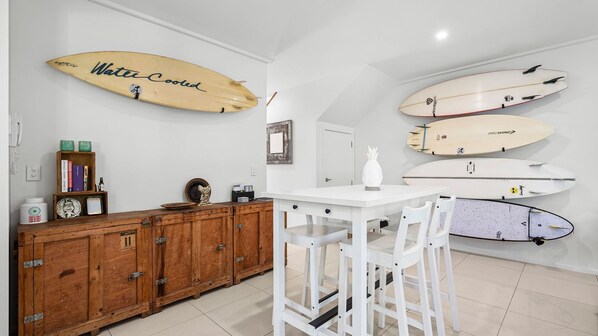 Una cocina con taburetes blancos, un mueble de madera y tablas de surf en la pared.