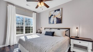 Een slaapkamer met een groot bed, een plafondventilator, een nachtkastje en een raam met gordijnen.
