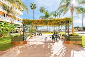 Ein Außenbereich mit Terrasse, einer mit Grün und orangefarbenen Blüten bewachsenen Pergola, umgeben von Palmen und einem Gebäude mit mehreren Balkonen.