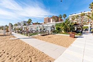 A beachfront hotel with a covered outdoor seating area, palm trees, and a sandy walkway.