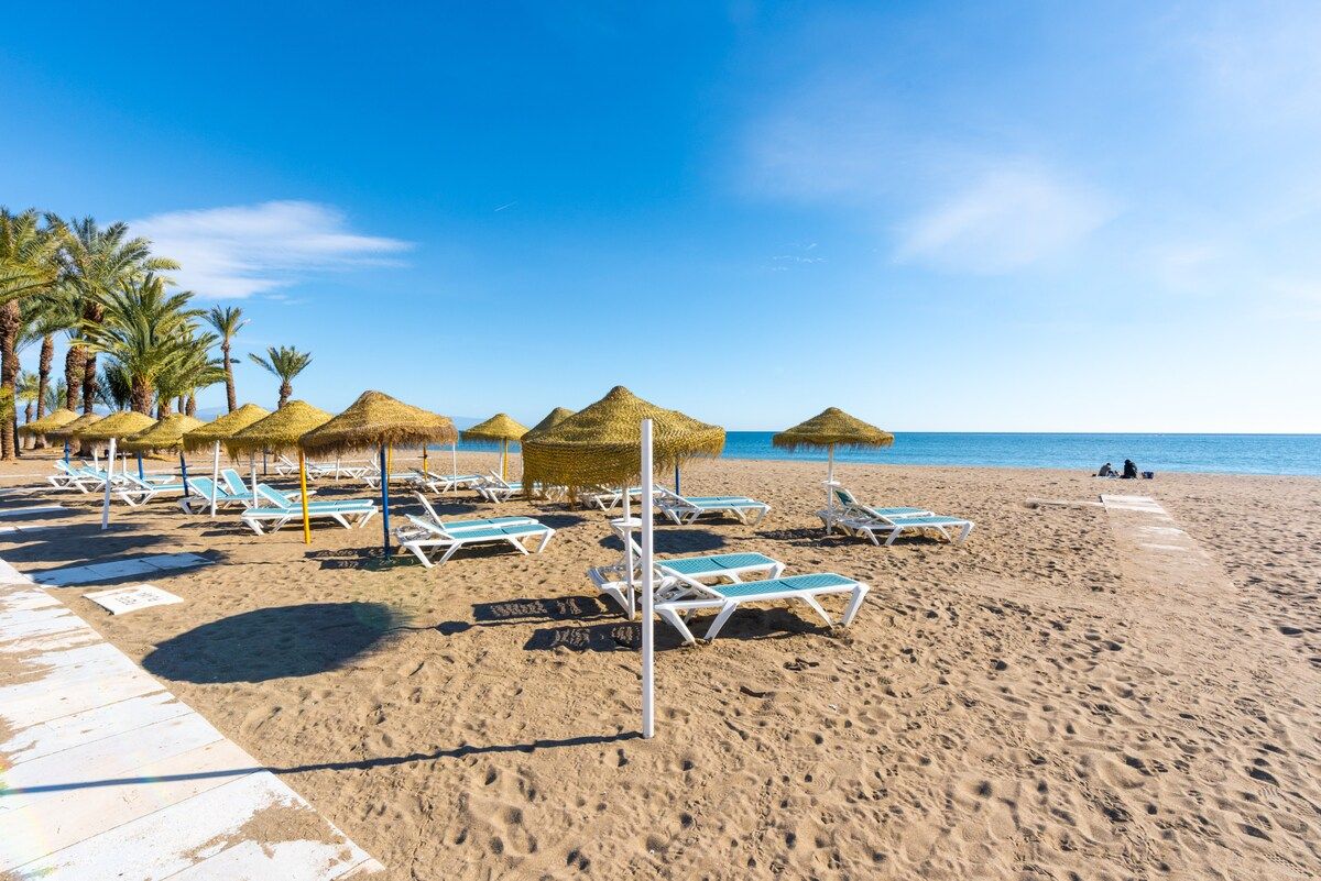 Une plage avec des chaises longues et des parasols en chaume, des palmiers et un ciel bleu dégagé.