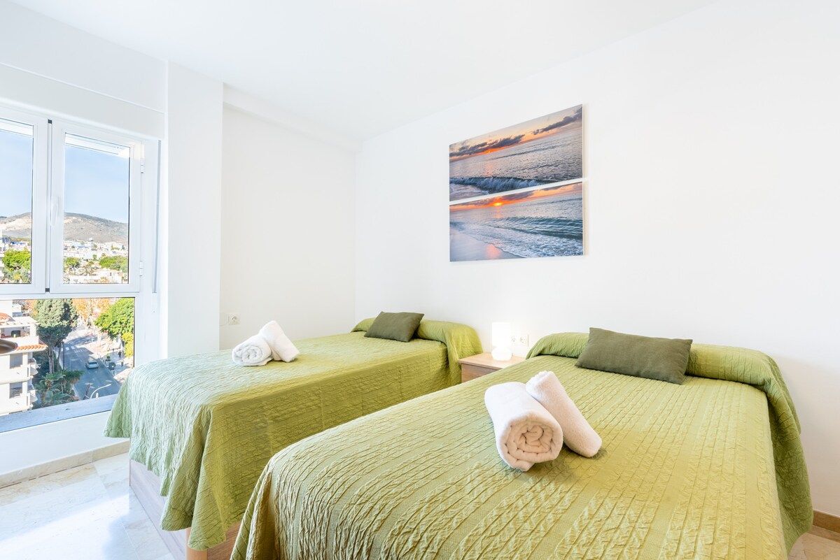 Una camera d'albergo con due letti, un quadro raffigurante un tramonto e una finestra vista edifici.