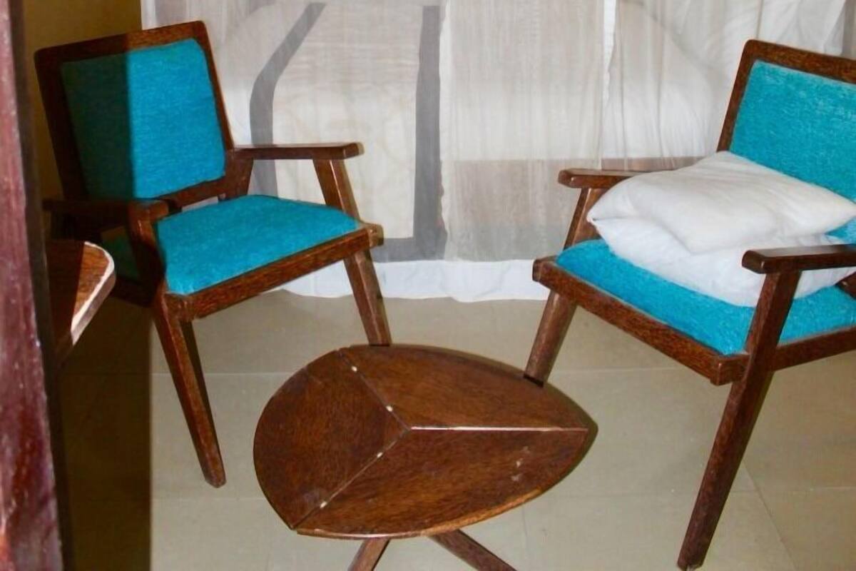 A room with a bed covered by a white canopy, two wooden chairs with blue cushions, and a small wooden table.
