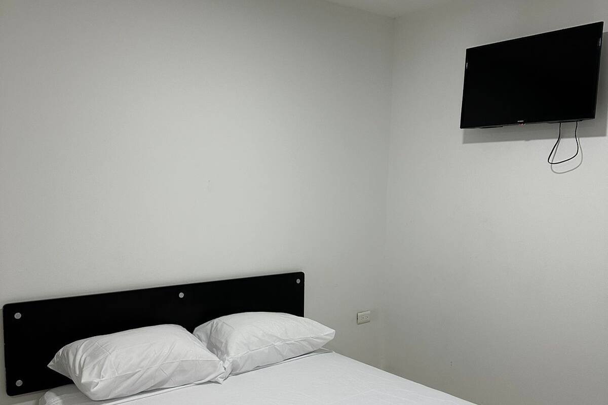 A single bed with white bedding in a room with white walls, a black headboard, and a flat-screen TV mounted on the wall.