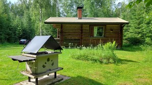 A rustic cabin with a chimney, surrounded by lush greenery and a barbecue grill in the foreground.