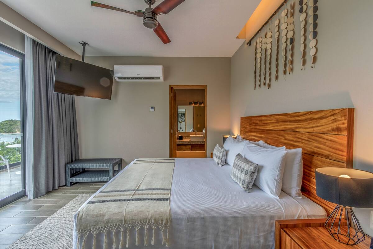 A modern bedroom with a large bed, a ceiling fan, a TV, and a balcony with a view.