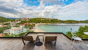 Un balcón con dos sillas y una mesa, con vista a una playa y un pueblo costero.