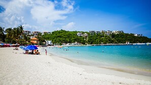 Una playa con cielo azul despejado, orilla arenosa y edificios al fondo.