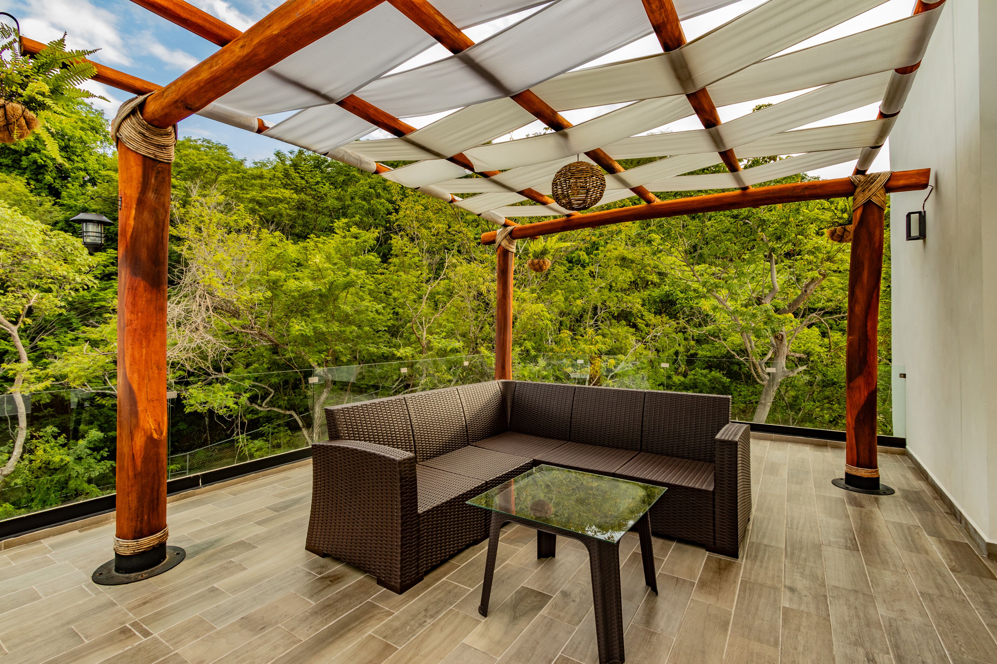 Un espace extérieur aménagé, abrité et doté d'un canapé en osier et d'une table en verre, entouré d'une pergola en bois et d'une végétation luxuriante.