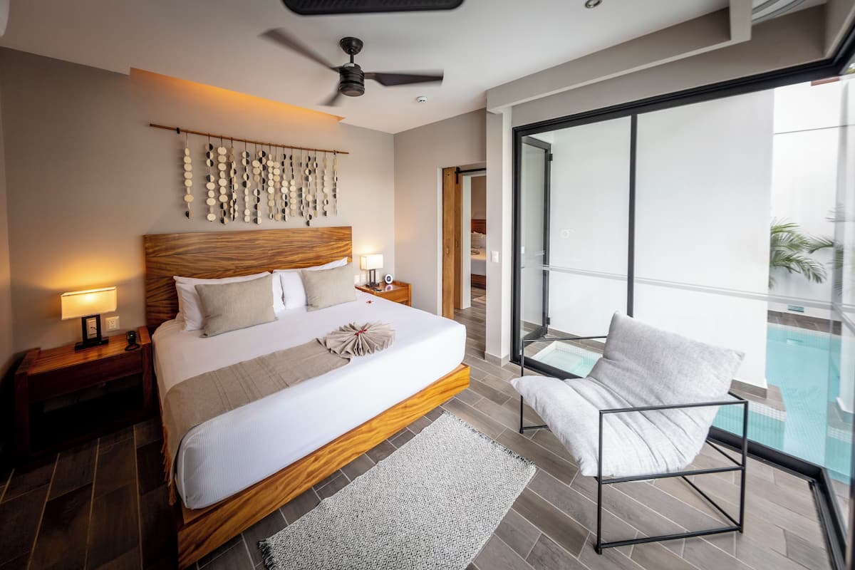 A modern hotel room with a large bed, a ceiling fan, a nightstand with a lamp, a view of a swimming pool through a glass door, and a chair by the pool.