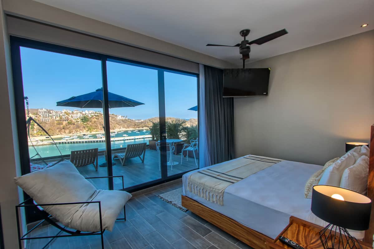A bedroom with a large window, a bed, a chair, a ceiling fan, and a TV mounted on the wall.