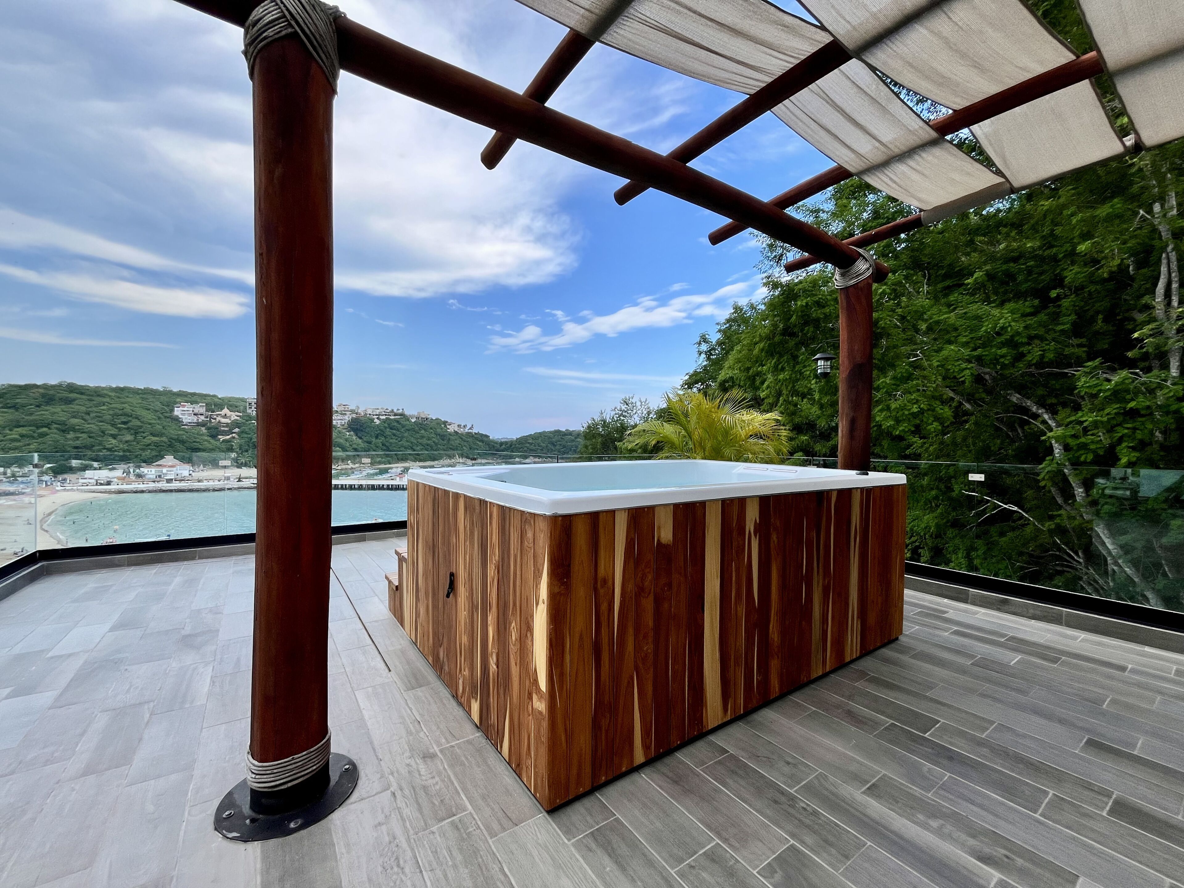A wooden hot tub on a balcony with a view of a beach and hills.