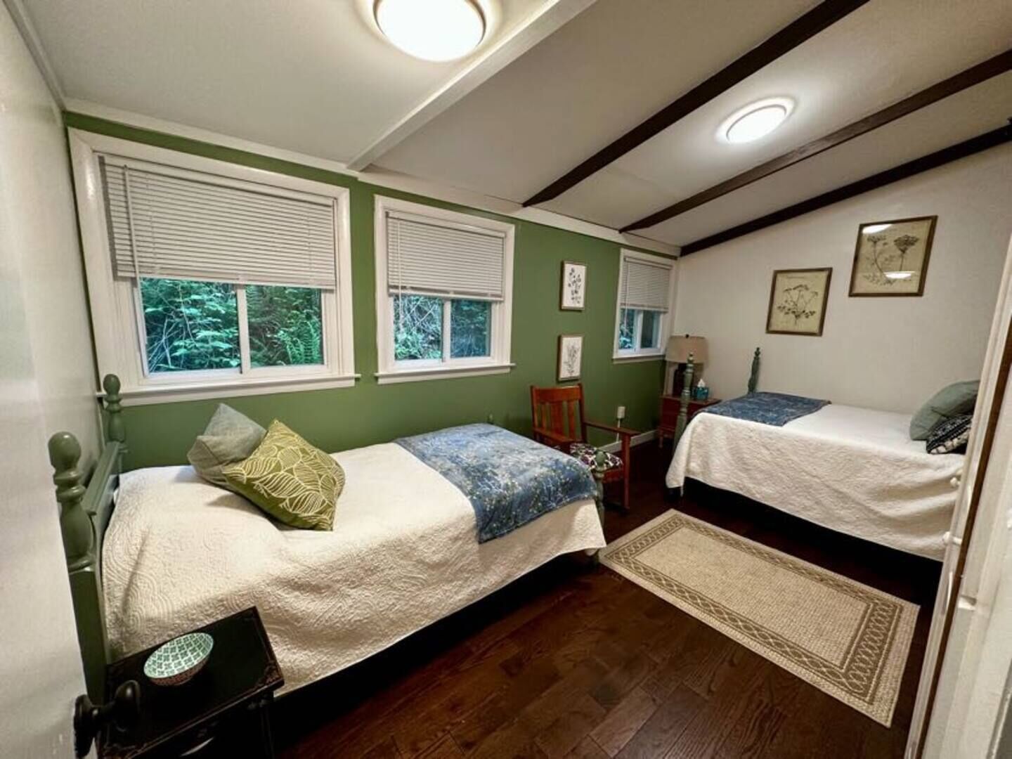 A room with two single beds, a small table, a chair, and a window with blinds.