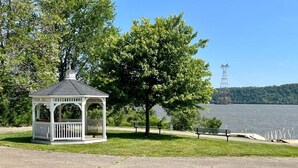 A gazebo with a roof and railings, surrounded by grass and trees, near a body of water with a distant power line tower. - Nature's Grove Cabin

 (Delta)