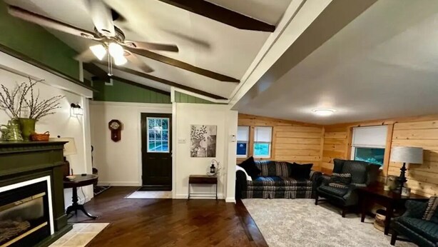 A spacious living room with hardwood flooring, a fireplace, a ceiling fan, and a large window. - Nature's Grove Cabin

 (Delta)