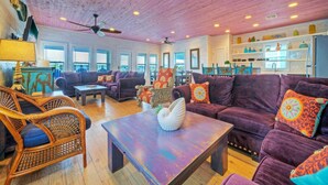 A spacious living room with a wooden ceiling, large windows, and a kitchen area with shelves. - Purple Pelican by Avantstay Waterfront Oasis (Port Aransas)