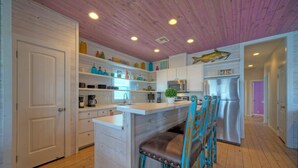 A kitchen with wooden floors, white cabinets, a stainless steel refrigerator, and a bar area with blue chairs. - Purple Pelican by Avantstay Waterfront Oasis (Port Aransas)