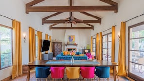 A dining area with a wooden table, colorful chairs, a fireplace, and a ceiling fan. - Luxe 7bdr Oasis w/ Pool, Putting Green, Game Room Villa Del Gallo by Avantstay (Indio)