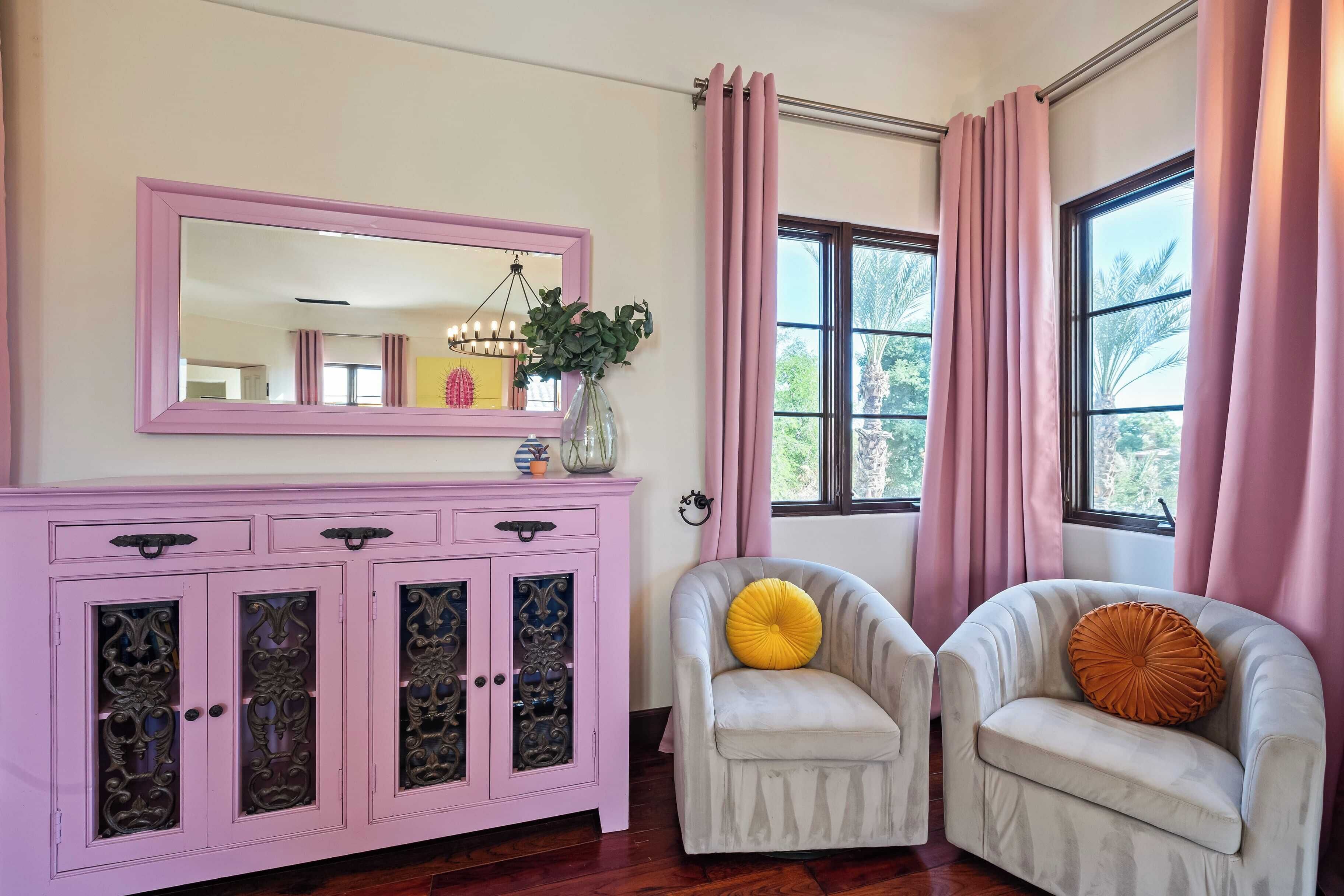 Une pièce avec un meuble rose, deux fauteuils, un miroir et une fenêtre avec des rideaux.