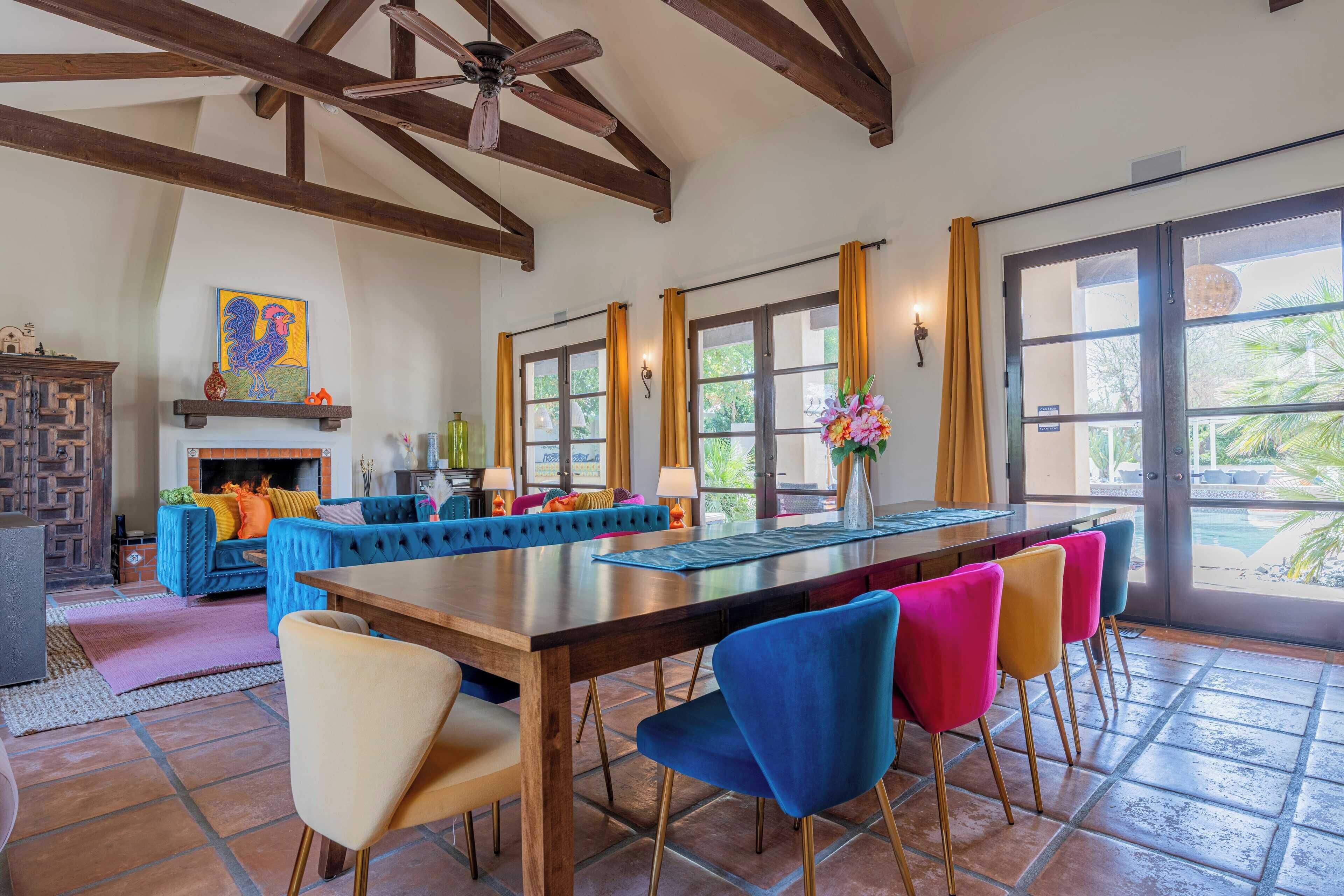 A spacious living area with a large wooden dining table, colorful chairs, a fireplace, and large windows with curtains.