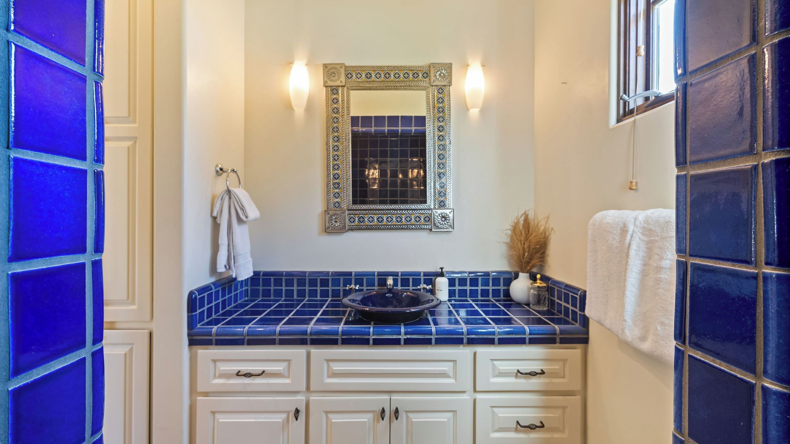 Un baño con lavamanos de azulejos azules, armarios blancos, un espejo con marco decorativo y un perchero para toallas.
