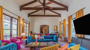 A spacious living room with a high ceiling, wooden beams, a large TV, and vibrant blue and pink sofas. - Luxe 7bdr Oasis w/ Pool, Putting Green, Game Room Villa Del Gallo by Avantstay (Indio)