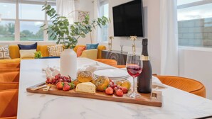 Ein Esstisch mit einer Auswahl an Käse, Trauben und einer Weinflasche, in einem modernen Wohnzimmer mit einer Couch und einem Fernseher.