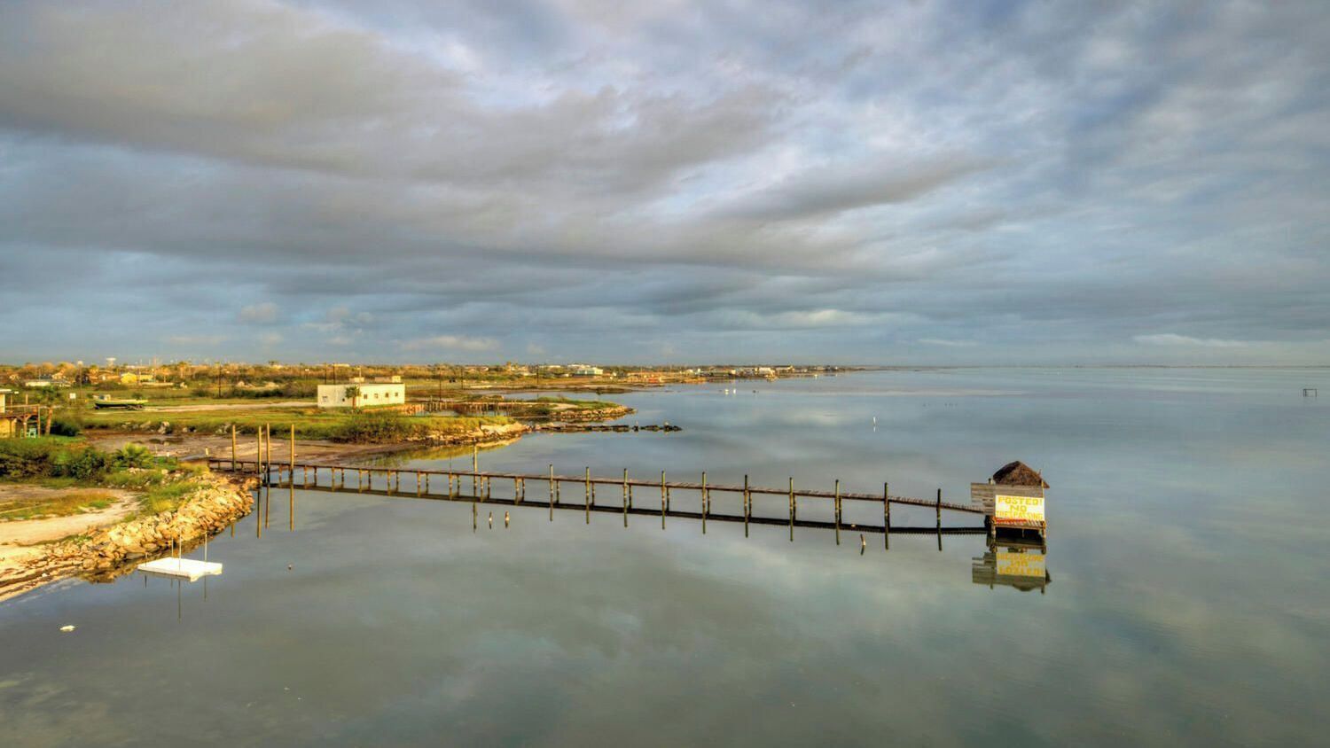A calm body of water with a wooden dock extending into it, a small hut at the end, and a distant shoreline with buildings.