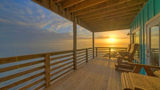 A wooden deck with a view of the sunset over the water, wooden chairs, and a glass door. - Waterfront w/ Bay Views, Shared Pool & Boat Dock Marlin Madness by Avantstay (Corpus Christi)