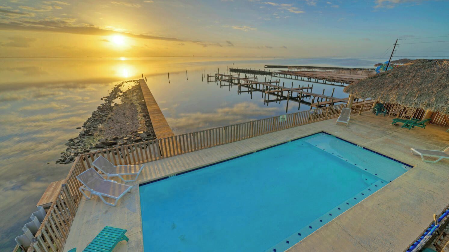 A swimming pool with a wooden deck, a thatched-roof structure, and a pier extending into the water.