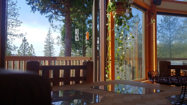 Un terrazzo in legno con tavolo e sedie in vetro, circondato da ampie finestre che si affacciano su alberi alti.