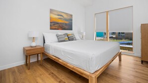 Ein Schlafzimmer mit einem hölzernen Bett, weißer Bettwäsche, einem Nachttisch mit Lampe, einem Sonnenuntergangsbild und einem großen Fenster mit Jalousien.