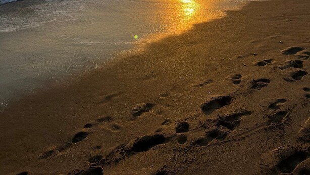 Ein Strand mit Fußabdrücken, die zum Wasser führen, Wellen und einem Sonnenuntergang.