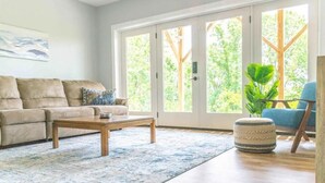 Un salon avec un canapé beige, une table basse en bois et un tapis à motifs. Une grande fenêtre offre une vue sur les arbres et on aperçoit une plante.
