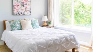 Une chambre avec une grande fenêtre, un lit agrémenté d’oreillers à motifs floraux, une table de chevet avec une lampe et une œuvre d’art florale encadrée accrochée au mur.