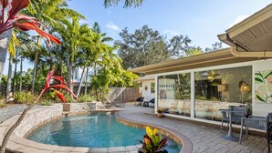 A swimming pool with a curved edge, surrounded by a stone deck, palm trees, and a glass building with a sign that reads "TOILETS".