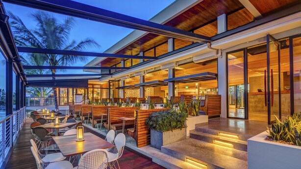 A modern outdoor dining area with wooden benches and tables, surrounded by glass railings and potted plants.