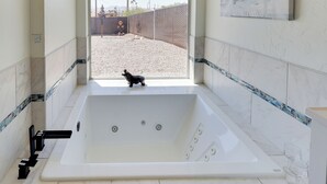 A modern bathroom with a large bathtub, a window with a view of a fenced outdoor area, and a small dog visible through the window. - Furnished Patio, Grill & Views: Lake Havasu Home (Lake Havasu City)