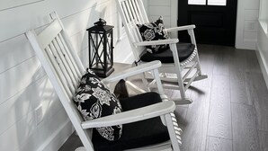Een veranda met twee witte schommelstoelen, zwarte kussens, een zwarte lantaarn en een bord met een raam.