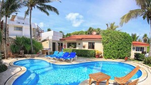 A swimming pool with lounge chairs and a small table, surrounded by palm trees and a building with a red roof. - Extraordinary Ocean Views - 2 bed / 2 bath and pool (Akumal)