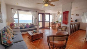 A spacious living area with a sofa, coffee table, and wicker chairs. There is a dining area with a table and chairs. The room has large windows with curtains, a ceiling fan, and a view of the ocean. - Extraordinary Ocean Views - 2 bed / 2 bath and pool (Akumal)