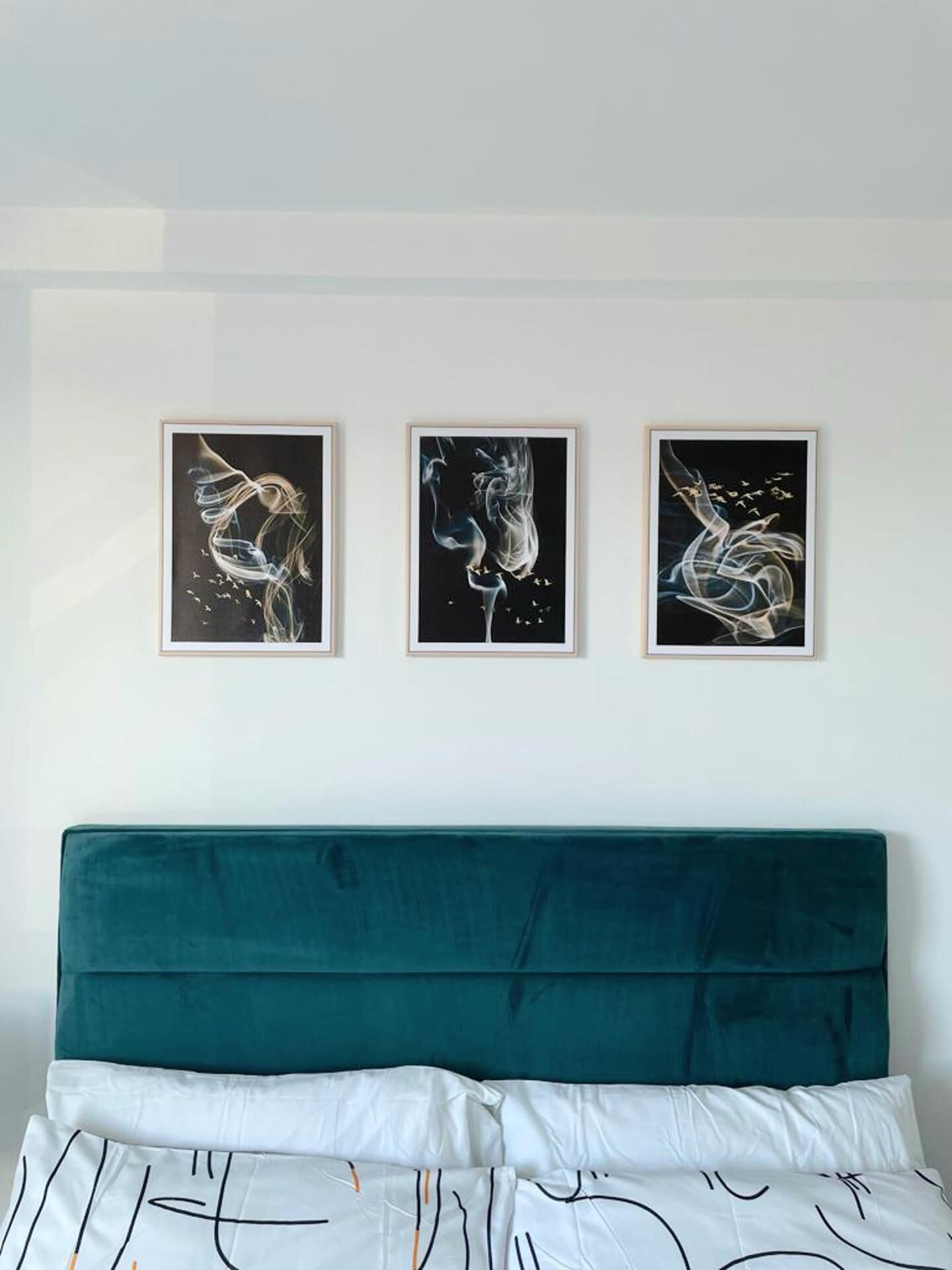 A bedroom with a teal headboard, white bedding, and three abstract art pieces on the wall.